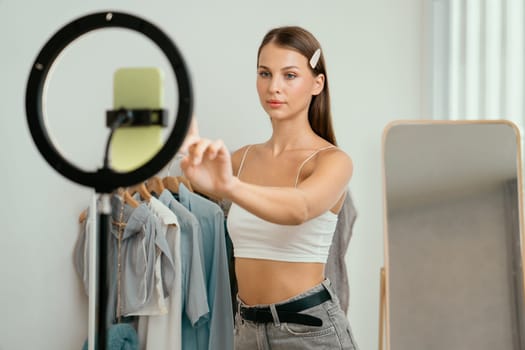 Young woman making fashion video content for social media. Blogger smiles to camera using selfie stick and light ring while making persuasive online clothing sell to audience or followers. Unveiling