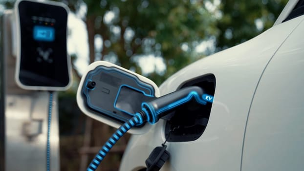 Electric car plugged in with charging station to recharge battery with electricity by EV charger cable in eco green park and greenery foliage. Future innovative ev car and energy sustainability.Peruse