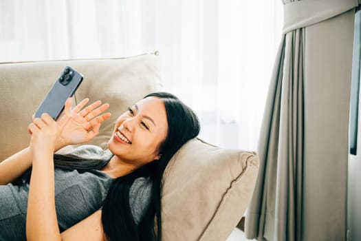 Happy young Asian woman on comfortable sofa texts on smartphone smiles. Enjoying relaxation chatting and shopping online. Modern technology for communication and connection at home.