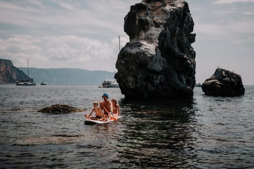 Father and his adorable little son and daughter sitting on stand up board having fun during summer beach vacation