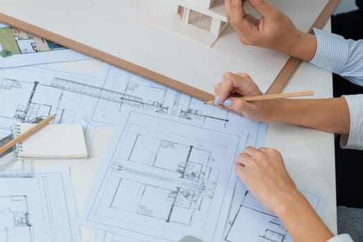 Top view image of professional beautiful young architect hand drafts blueprint while asian cooperative coworker measure house model on table with house model and blueprint scatter around. Immaculate.