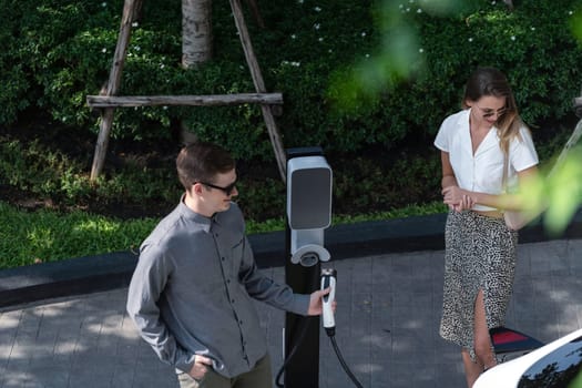 Young couple travel with EV electric car charging in green sustainable city outdoor garden in summer shows urban sustainability lifestyle by green clean rechargeable energy of electric vehicle innards