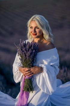 Blonde woman poses in lavender field at sunset. Happy woman in white dress holds lavender bouquet. Aromatherapy concept, lavender oil, photo session in lavender.