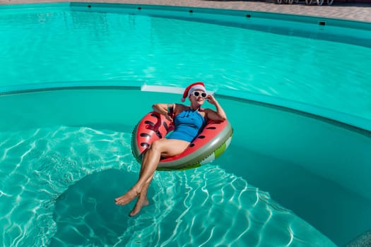 A happy woman in a blue bikini, a red and white Santa hat and sunglasses poses in the pool in an inflatable circle with a watermelon pattern, holding a glass of wine in her hands. Christmas holidays concept