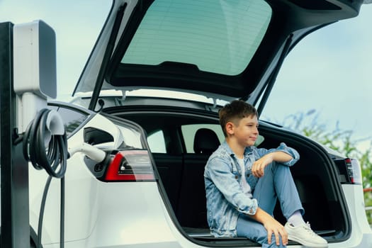 Little boy sitting on car trunk while recharging eco-friendly electric car from EV charging station. EV car road trip travel concept for alternative transportation powered sustainable energy.Perpetual