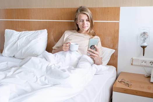 Woman using smartphone and drinking coffee lying in bed at hotel room