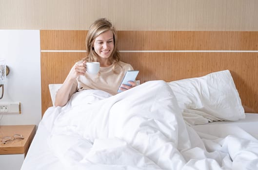 Woman using smartphone and drinking coffee lying in bed at hotel room