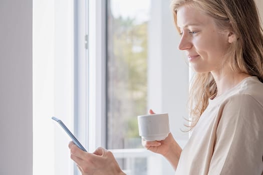 woman standing beside a window holding mobile phone drinking coffee