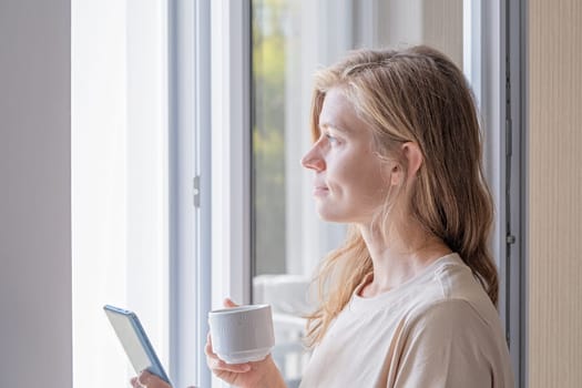woman standing beside a window holding mobile phone drinking coffee