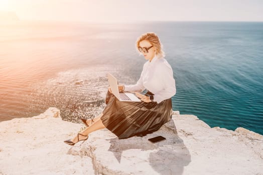 Successful business woman in yellow hat working on laptop by the sea. Pretty lady typing on computer at summer day outdoors. Freelance, travel and holidays concept.