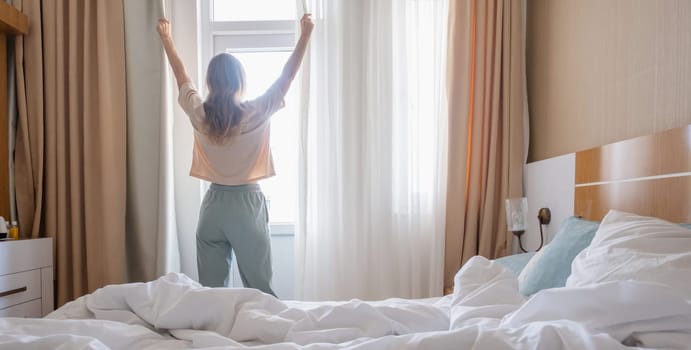 Woman stretching standing by window after wake up, back view, sun shines from the window