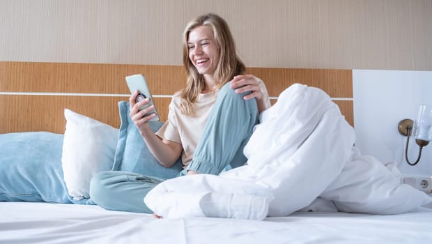 Woman using smartphone and laughing sitting in bed at hotel room