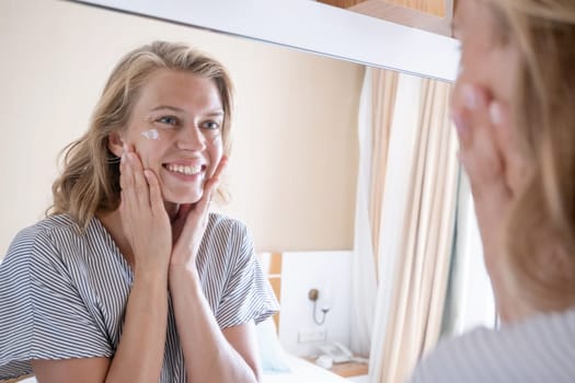 Beautiful woman making up by the mirror in hotel room , applying face cream