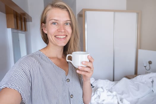 Vacation concept. Happy woman showing her hotel room taking selfie holding cup of coffee