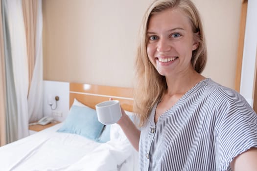 Vacation concept. Happy woman showing her hotel room taking selfie holding cup of coffee