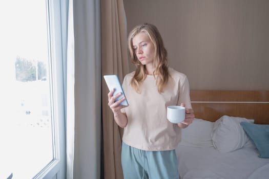 woman standing beside a window holding mobile phone drinking coffee