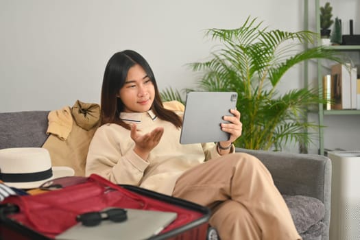 Smiling Asian woman having video call on digital tablet while preparing travel suitcase at home.