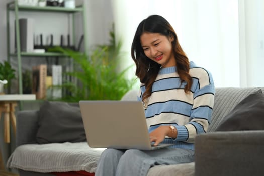 Smiling young woman relaxing on couch at home and using laptop. People and technology concept.