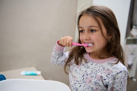 Caucasian little cute child girl brushing her teeth in the gray bathroom. Copy advertising space for text. Healthy teeth. Oral care and dental hygiene. Healthy habits to prevent caries and tooth decay