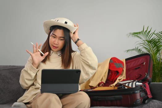 Young woman having video call on digital tablet while preparation for a vacation trip at home.