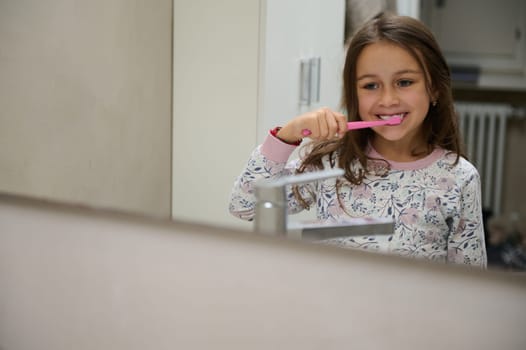 Adorable Caucasian little kid girl in pajamas, looking at her mirror reflection while brushing teeth in the the bathroom. Healthy lifestyle. Healthy habits. Dental care and hygiene concept. Copy space