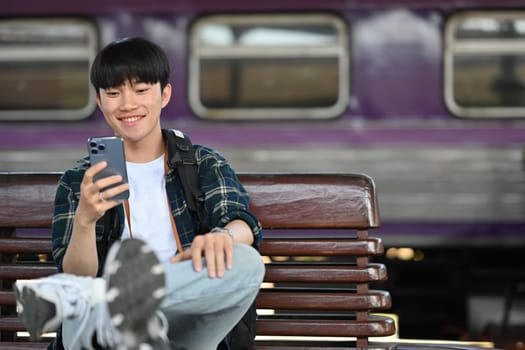 Happy young Asian man traveling by himself with backpack, using mobile phone at train station.