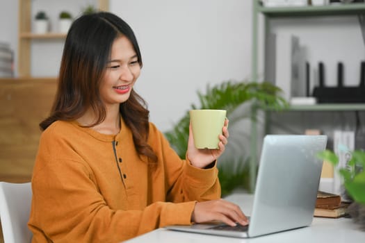 Young female freelancer holding cup of coffee and working on laptop at home.