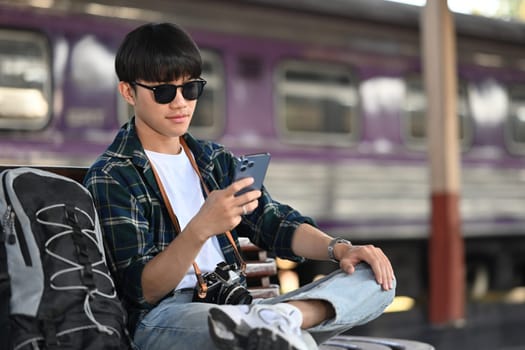 Male tourist texting message on mobile phone, sitting on bench at railway station.