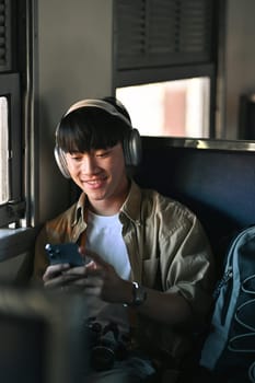 Happy man in headphones using mobile phone while sitting near the window in train during the trip