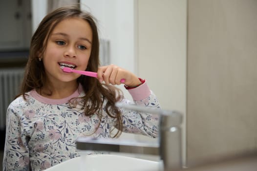 Close-up authentic portrait of a Caucasian cute child girl brushing teeth, admiring herself in the bathroom mirror. Dental hygiene, oral care and healthy lifestyle concept