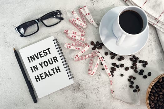 INVEST IN YOUR HEALTH text on a notebook with cup of coffee, coffee bean and measurement tap on a desk.