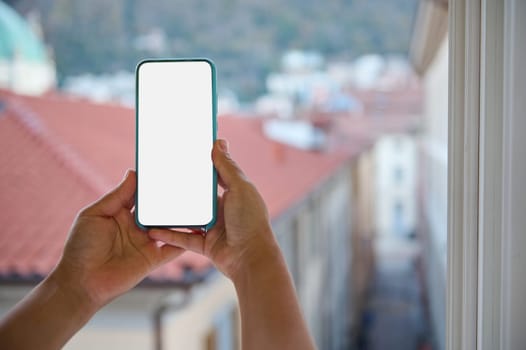Close-up hands hold smartphone with white blank mockup screen, against backdrop of beautiful city with houses with red roof tiles, photographing cityscape in the morning. Copy space for for mobile app
