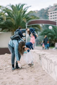 Young woman in gloves bending over collects garbage from the beach into a bag. High quality photo