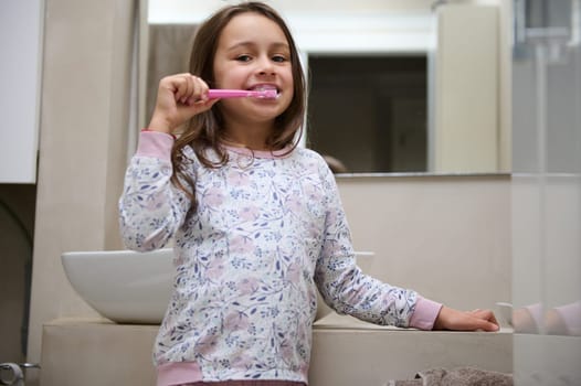 Little kid girl in pajamas, brushing her teeth, standing in the gray minimalist home bathroom, smiling looking at camera. Dental care. Oral hygiene. Healthy lifestyle. Prevent of caries. Copy ad space