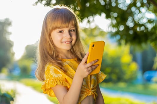 Preteen child kid using smartphone typing text answering messages chatting online looking mobile screen social media app, playing game. Pretty blonde girl standing on city sunset park street, outdoors