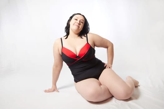 Portrait of attractive dreamy thick woman in red black swimsuit posing on white background. Body positive, photoshoot, selfie. Funny plus size model