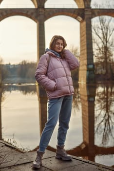 A young woman standing at the shore looking at the river in autumn sunny day. Street view, copy space for text, travel photo. Happy tourist woman on the bank of the river in autumn in warm clothes. Tourists enjoy their vacation, winter season. Romantic look and travel concept. A joyful mood in a Caucasian girl. Winter Wonderland: Enchanting Girl by the Riverside in Autumn.