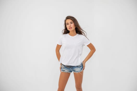 Beautiful brunette girl in a white T-shirt and denim shorts posing on a white background. High quality photo