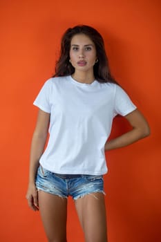 Beautiful brunette girl in a white T-shirt and denim shorts posing on an orange background. High quality photo
