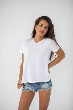 Beautiful brunette girl in a white T-shirt and denim shorts posing on a white background. High quality photo