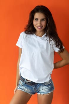 Beautiful brunette girl in a white T-shirt and denim shorts posing on an orange background. High quality photo