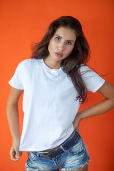 Beautiful brunette girl in a white T-shirt and denim shorts posing on an orange background. High quality photo