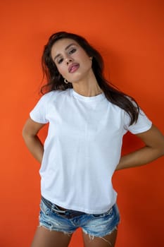 Beautiful brunette girl in a white T-shirt and denim shorts posing on an orange background. High quality photo