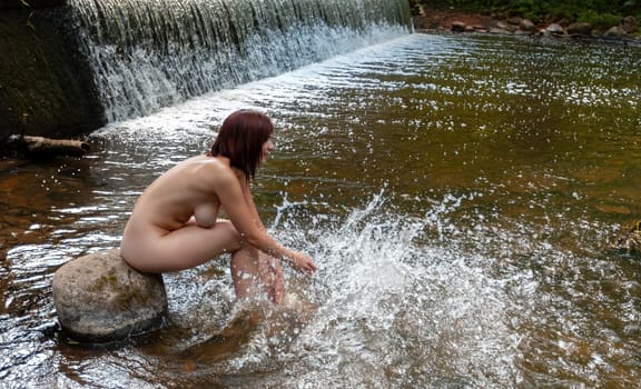 Young nude woman enjoying the fresh coolness near a forest stream with a waterfall