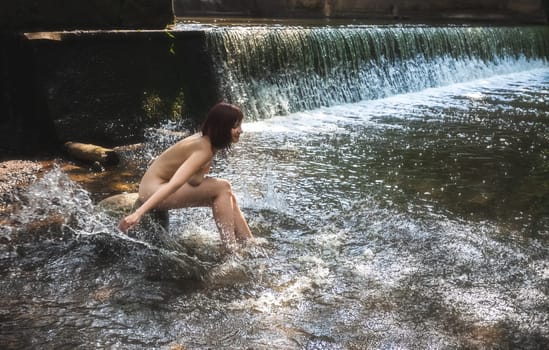 Young nude woman enjoying the fresh coolness near a forest stream with a waterfall