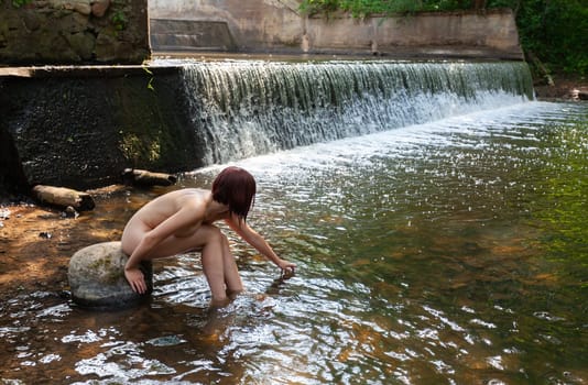 Young nude woman enjoying the fresh coolness near a forest stream with a waterfall