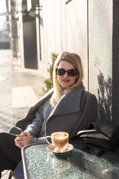 Portrait of tanned lady with cup of latte on foreground. Smiling woman in sunglasses during breakfast in cafe. High quality photo