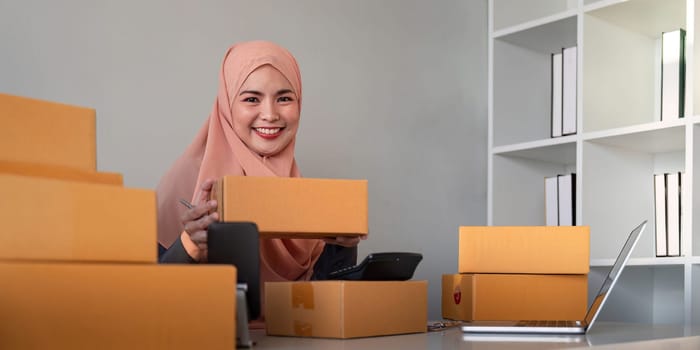 Muslim women selling online at home with box. Selling online with box to accept order from customer. SME business idea. Parcel delivery. muslim woman working smartphone and laptop at home.