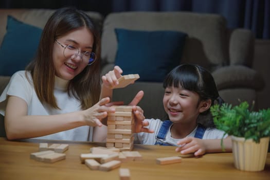 Asian young mother playing game in wood block with little daughter in home living room at night, Smiling woman help teach preschooler kid play build constructor tower of wooden blocks, family funny