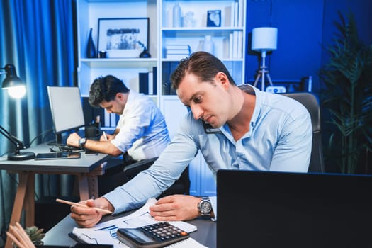 Serious businessman calling on phone with customer to create new project at night time, coworker writing document work sheet on desk. Concept of working on neon blue light modern office. Sellable.